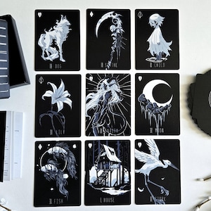 May include: A black tarot card deck with illustrations in white. The deck includes a box, a booklet, and a deck of cards. The cards feature images of a wolf, a scythe, a child, a lily, a woman, a moon, a fish, a house, and a crane.