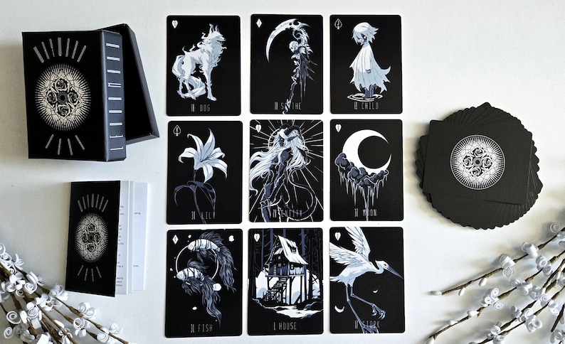 May include: A black tarot card deck with illustrations in white. The deck includes a box, a booklet, and a deck of cards. The cards feature images of a wolf, a scythe, a child, a lily, a woman, a moon, a fish, a house, and a crane.