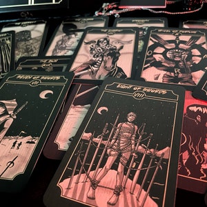 May include: A collection of tarot cards with black and gold designs. The cards feature illustrations of figures and symbols, including swords, moons, and stars. The cards are arranged on a dark surface, with some cards displaying titles like "Eight of Swords" and "Seven of Swords."