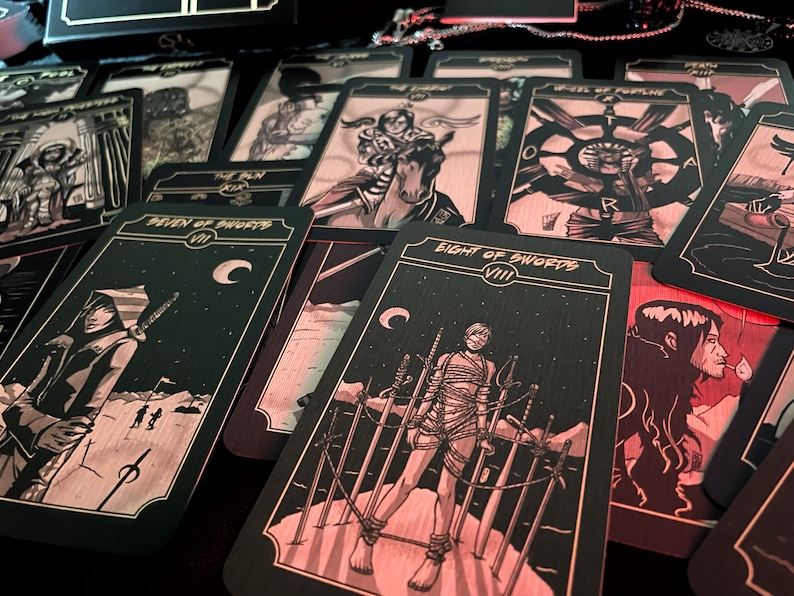 May include: A collection of tarot cards with black and gold designs. The cards feature illustrations of figures and symbols, including swords, moons, and stars. The cards are arranged on a dark surface, with some cards displaying titles like "Eight of Swords" and "Seven of Swords."