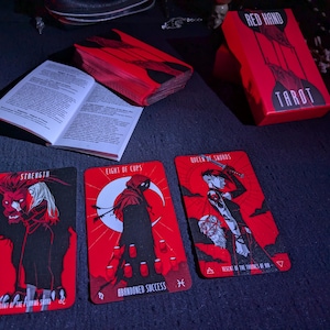 May include: Three tarot cards from the Red Hand Tarot deck. The cards are Strength, Eight of Cups, and Queen of Swords. The cards are red with black illustrations.