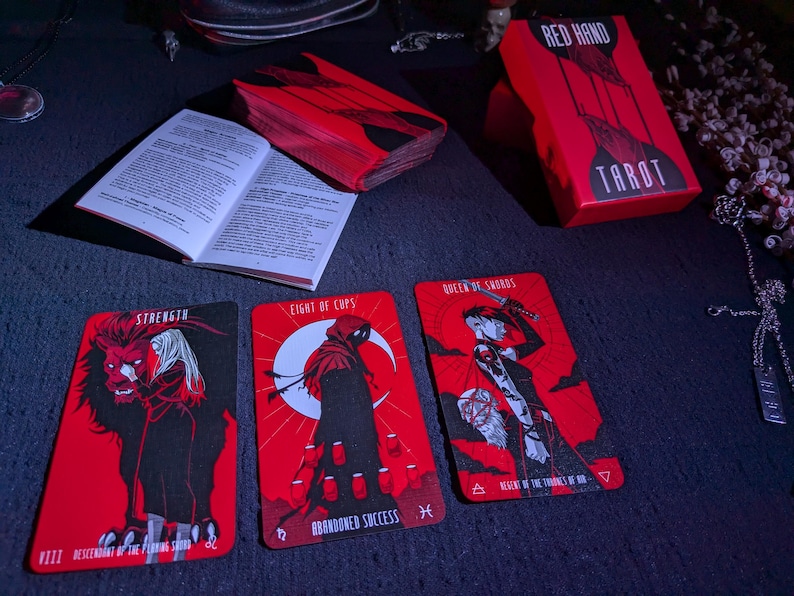 May include: Three tarot cards from the Red Hand Tarot deck. The cards are Strength, Eight of Cups, and Queen of Swords. The cards are red with black illustrations.