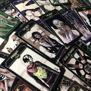 May include: A collection of tarot cards with black borders and gold accents. Each card features unique illustrations and titles such as "Strength," "The Devil," and "Wheel of Fortune." The cards are arranged in a scattered layout, showcasing their detailed artwork.