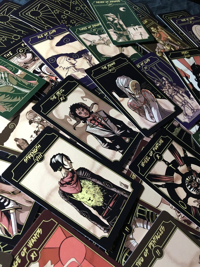 May include: A collection of tarot cards with black borders and gold accents. Each card features unique illustrations and titles such as "Strength," "The Devil," and "Wheel of Fortune." The cards are arranged in a scattered layout, showcasing their detailed artwork.