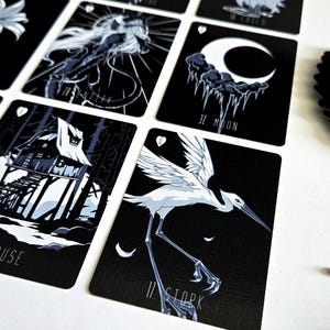 May include: A deck of tarot cards with black backgrounds and white illustrations. The cards feature images of a lily, a jellyfish, a crescent moon, a house, and a stork. A leaf-shaped card holder is visible.