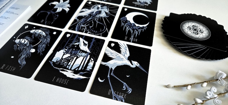 May include: A deck of tarot cards with black backgrounds and white illustrations. The cards feature images of a lily, a jellyfish, a crescent moon, a house, and a stork. A leaf-shaped card holder is visible.