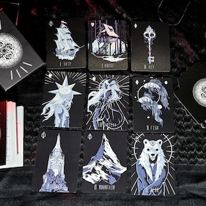May include: A deck of tarot cards with dark illustrations. The cards feature images of a ship, house, key, and other mystical figures. The card backs and box have a floral design. The cards are black with white and blue accents.
