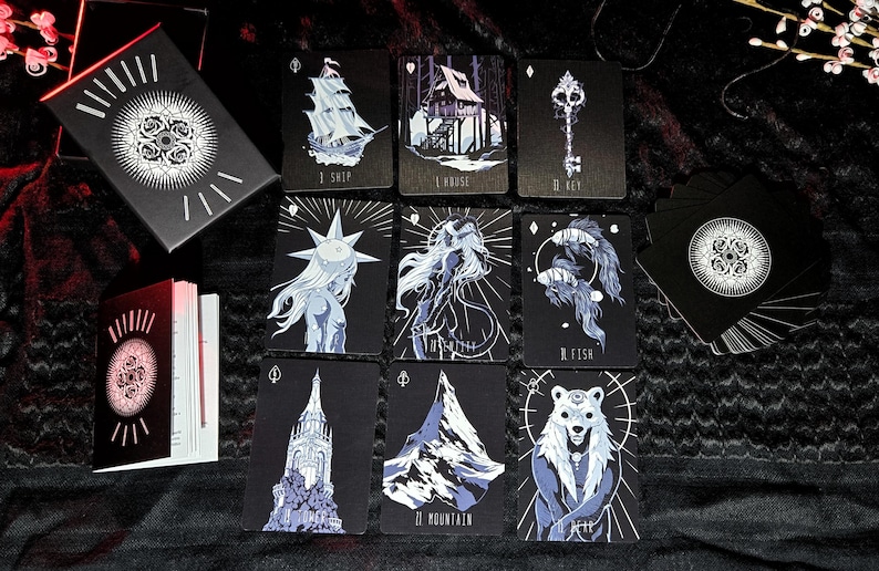 May include: A deck of tarot cards with dark illustrations. The cards feature images of a ship, house, key, and other mystical figures. The card backs and box have a floral design. The cards are black with white and blue accents.