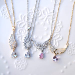 May include: Four necklaces with wing pendants. Two are gold-coloured, and two are silver-coloured. Each pendant features a wing design with clear or pink gemstones. The necklaces are displayed on a white surface.