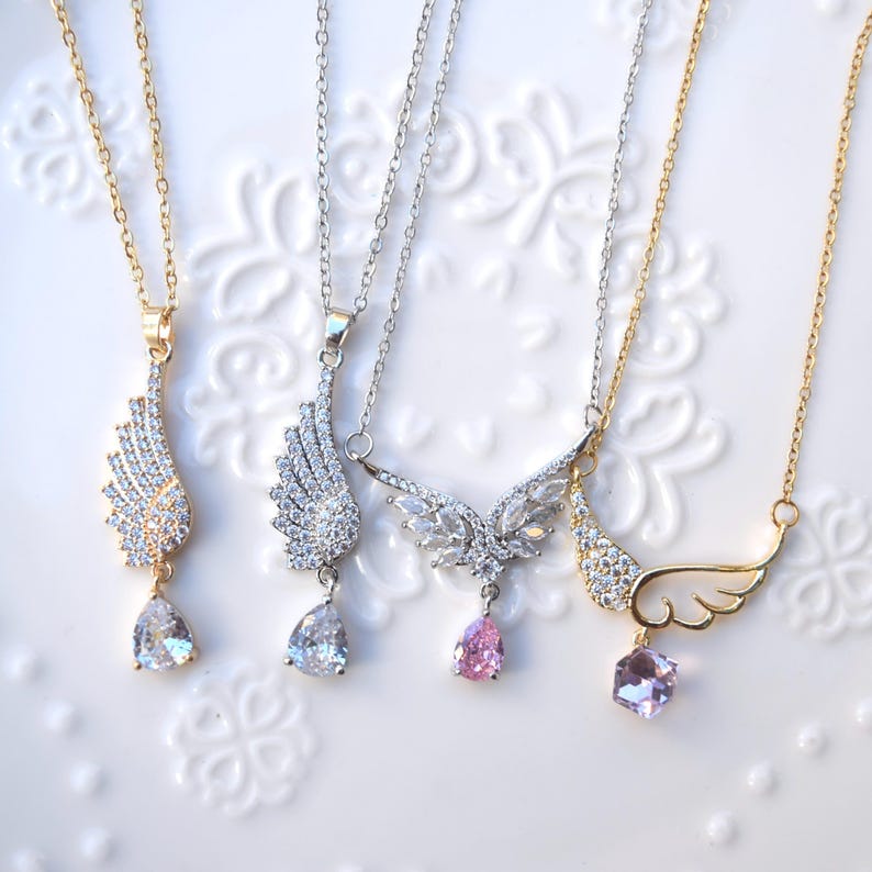 May include: Four necklaces with wing pendants. Two are gold-coloured, and two are silver-coloured. Each pendant features a wing design with clear or pink gemstones. The necklaces are displayed on a white surface.