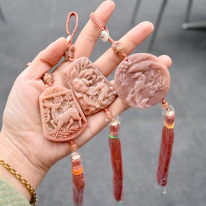 May include: A collection of pink-toned carved pendants with intricate designs, including a deer, a rabbit, and a bird. Each pendant is attached to a red tassel and a woven cord, creating a decorative accessory.