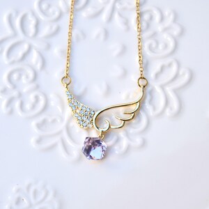 May include: A gold-coloured necklace with a wing pendant. One wing is embellished with small, clear crystals, the other is a solid wing shape. A faceted, light purple gemstone hangs below. The necklace is displayed on a white, patterned surface.