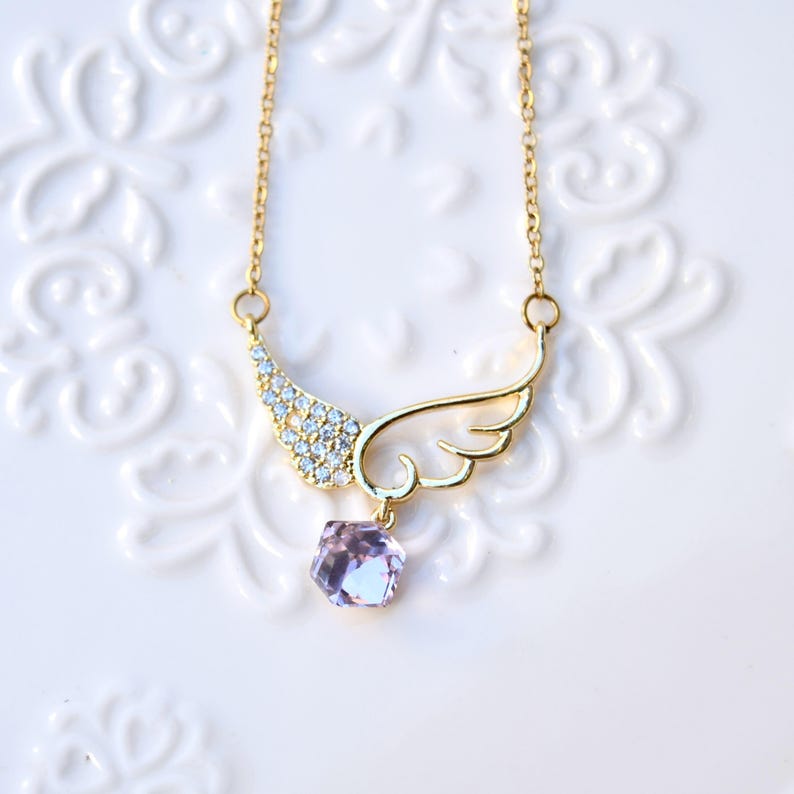 May include: A gold-coloured necklace with a wing pendant. One wing is embellished with small, clear crystals, the other is a solid wing shape. A faceted, light purple gemstone hangs below. The necklace is displayed on a white, patterned surface.