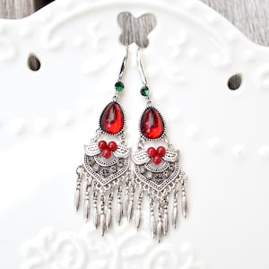 May include: A pair of silver-toned dangle earrings. Each earring features a teardrop-shaped red gem, a cluster of red beads, and a decorative silver filigree design with dangling silver accents. Green gems are at the top.