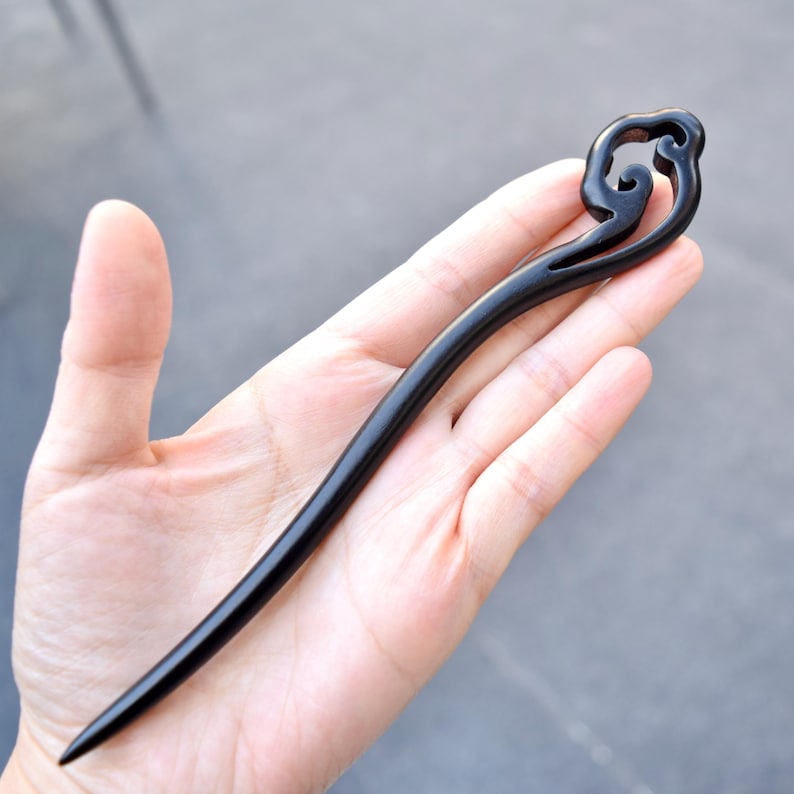 Pu&ograve; includere: Un fermacapelli in legno nero con un design decorativo a spirale sulla parte superiore. L'accessorio per capelli &egrave; tenuto in una mano, mostrando la sua superficie liscia e lucida e la sua forma elegante. Questo fermacapelli &egrave; un accessorio elegante.