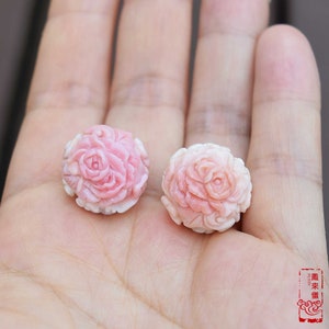 Peut inclure: Deux perles roses claires en forme de rose sculptée. Les perles sont rondes avec des pétales détaillés et une surface légèrement texturée. Les perles sont tenues dans la main d'une personne, mettant en valeur le design floral.