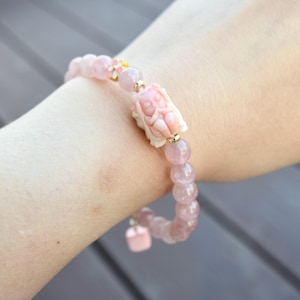 May include: A pink rose quartz bracelet with a carved ivory-colored centerpiece. The bracelet features round beads and a small pink square charm. Accents include gold-colored metal and a small floral design.
