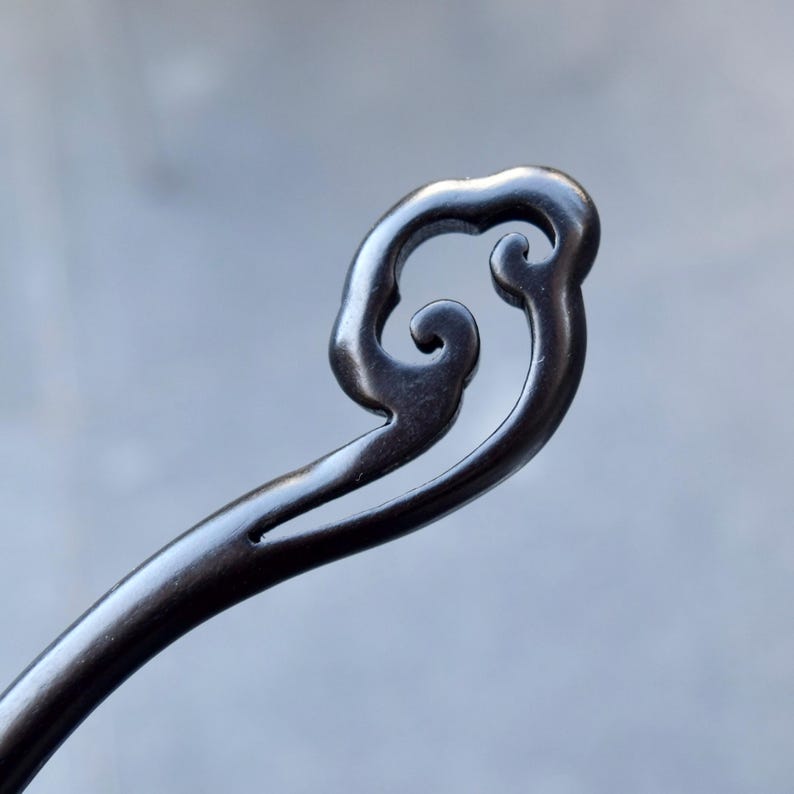 Pu&ograve; includere: Un fermaglio per capelli scuro e ornato con un design a spirale a forma di nuvola nella parte superiore. L'accessorio per capelli &egrave; realizzato in un materiale scuro, forse legno o metallo, e ha una finitura liscia e lucida. Il design &egrave; dettagliato ed elegante.
