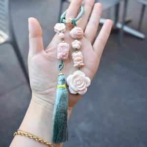 May include: A decorative pendant with carved pink and white beads, including a rose, animal figures, and floral shapes. The pendant is strung on a green cord with a matching tassel. A gold-coloured accent piece is at the top.