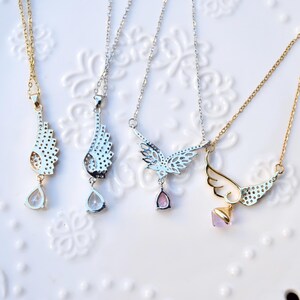 May include: Four necklaces with wing-shaped pendants. Two have gold chains and pendants, and two have silver chains and pendants. Each pendant has a different design, with clear or pink teardrop-shaped or square-shaped gemstones.