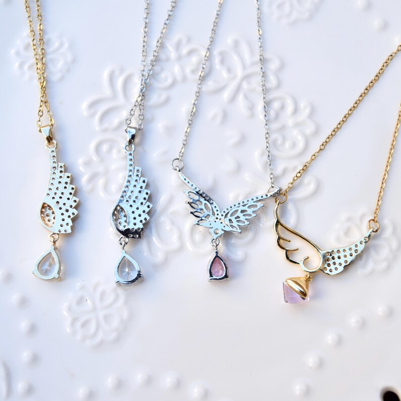 May include: Four necklaces with wing-shaped pendants. Two have gold chains and pendants, and two have silver chains and pendants. Each pendant has a different design, with clear or pink teardrop-shaped or square-shaped gemstones.
