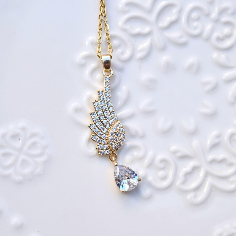 May include: A gold-tone necklace featuring a wing-shaped pendant adorned with clear crystals. The wing design is detailed with multiple rows of crystals, and a teardrop-shaped crystal hangs below. The necklace is displayed on a white surface.
