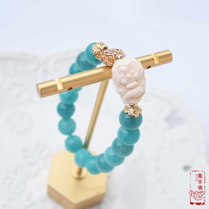 May include: A turquoise green beaded bracelet with a gold charm featuring a carved white figure. The bracelet is displayed on a gold stand.