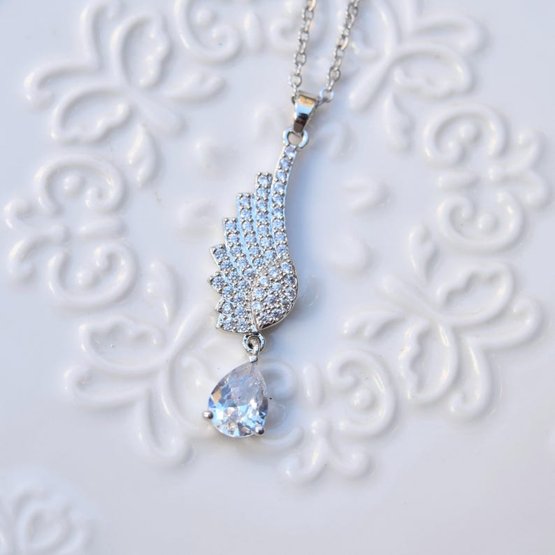 May include: A silver necklace featuring a wing-shaped pendant adorned with small, clear crystals. A clear, teardrop-shaped crystal dangles below the wing. The necklace is presented on a white, patterned surface.