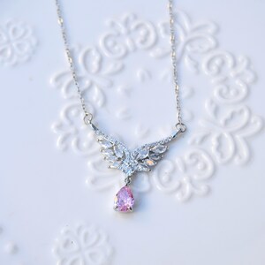May include: A silver necklace featuring a wing-shaped pendant set with clear crystals and a pink, pear-cut gemstone. The necklace is presented on a white, patterned surface, highlighting its intricate design and sparkle.