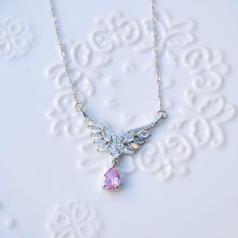 May include: A silver necklace featuring a wing-shaped pendant set with clear crystals and a pink, pear-cut gemstone. The necklace is presented on a white, patterned surface, highlighting its intricate design and sparkle.