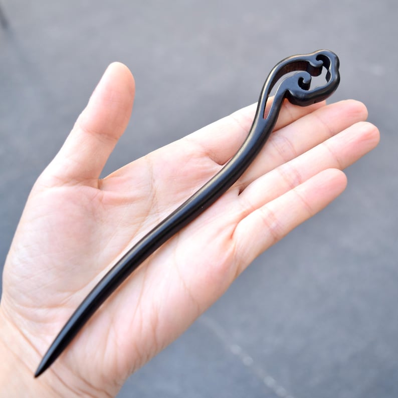 Pu&ograve; includere: Un fermacapelli in legno nero con un design curvo. La parte superiore presenta un motivo a spirale decorativo, mentre il bastoncino si assottiglia fino a una punta. L'accessorio per capelli &egrave; tenuto in una mano, mostrando la sua finitura elegante e lucida.