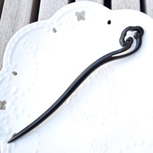 Pu&ograve; includere: Un fermacapelli in legno marrone scuro con un design curvo e fluente nella parte superiore. Il bastoncino &egrave; lungo e sottile, con un'estremit&agrave; appuntita. L'accessorio per capelli &egrave; posto su un piatto bianco decorativo con motivi floreali in rilievo.