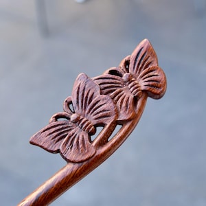 May include: A wooden hair stick with a carved butterfly design. The hair accessory is made of dark brown wood and features two detailed butterflies at the top. The butterflies have intricate wing patterns and are attached to a curved stick.
