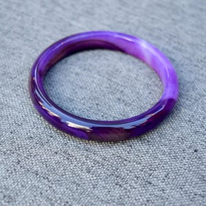 May include: A smooth, circular bangle bracelet crafted from polished purple agate stone. The material features translucent and opaque bands of deep violet and lighter lavender hues, creating a continuous, glossy, and rounded surface.