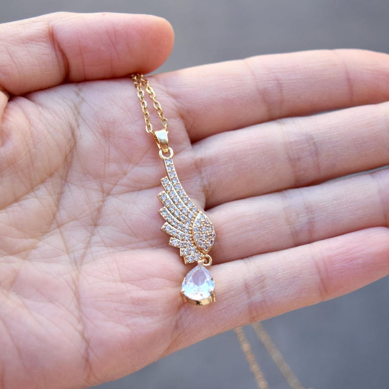 May include: A gold necklace featuring a wing-shaped pendant adorned with small, clear crystals. A teardrop-shaped, clear crystal hangs below the wing. The necklace is held in a hand. The chain is gold-colored.