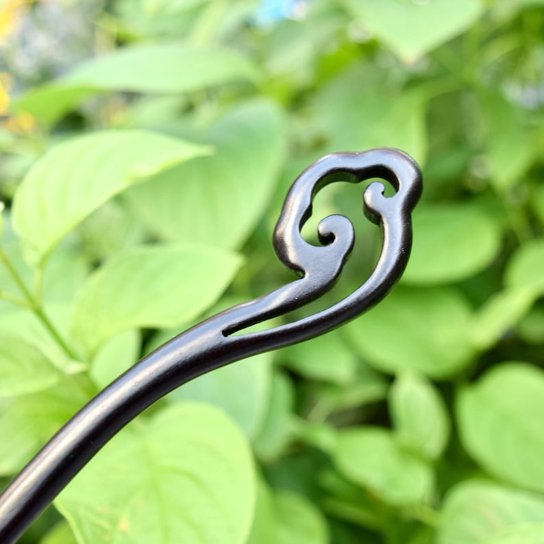 Pu&ograve; includere: Un fermacapelli in legno marrone scuro con un design a spirale decorativo sulla parte superiore. L'accessorio per capelli &egrave; posto su uno sfondo di fogliame verde, evidenziando la sua forma elegante e la sua maestria.