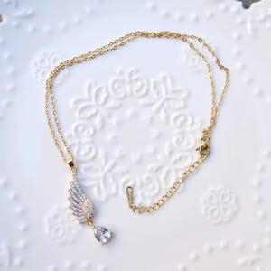 May include: Gold-coloured necklace with a wing-shaped pendant set with clear crystals and a teardrop-shaped crystal. The necklace is displayed on a white, patterned surface.