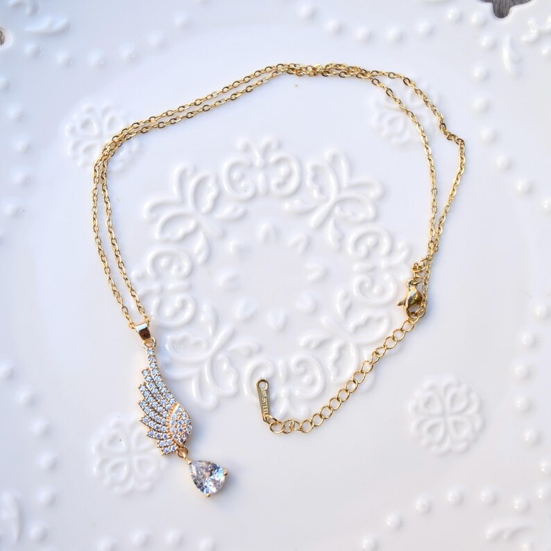 May include: Gold-coloured necklace with a wing-shaped pendant set with clear crystals and a teardrop-shaped crystal. The necklace is displayed on a white, patterned surface.