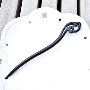 Pu&ograve; includere: Un fermacapelli nero con un design decorativo a forma di nuvola nella parte superiore. Il bastoncino &egrave; lungo e sottile, con una finitura liscia e lucida. L'accessorio per capelli &egrave; posto su un piatto bianco ornato.