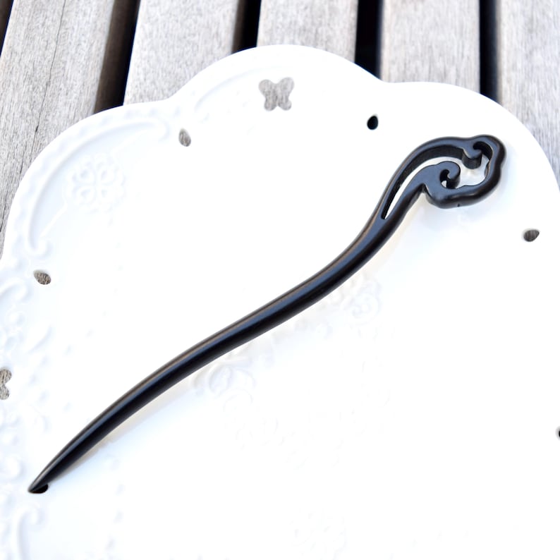 Pu&ograve; includere: Un fermacapelli nero con un design decorativo a forma di nuvola nella parte superiore. Il bastoncino &egrave; lungo e sottile, con una finitura liscia e lucida. L'accessorio per capelli &egrave; posto su un piatto bianco ornato.