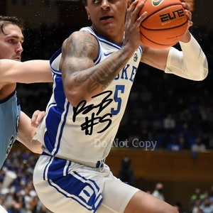 May include: A basketball player wearing a white and blue jersey with the number "5" on the front. The player is holding a basketball with the word "ELITE" on it. The player is in mid-air and has a determined look on his face. The player's jersey has the word "DUKE" on the front.
