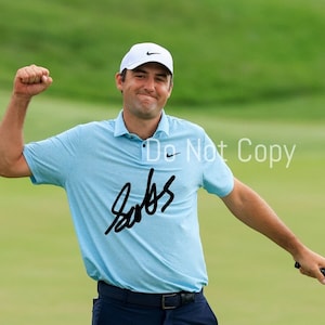 May include: A golfer wearing a light blue polo shirt with a white Nike logo and a white hat with a Nike logo. The golfer is raising his right arm in the air and holding a golf club in his left hand. The golfer's signature is written in black ink on the shirt. The signature says "Sabbatini".