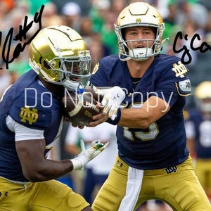 May include: Two football players in navy blue and gold uniforms are holding a football. Both players wear gold helmets and white gloves. The image has signatures in the top corners. The background is blurred with green and yellow.