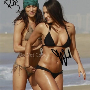 May include: Two women in bikinis on a beach. One woman is wearing a black bikini and the other is wearing a brown bikini. The woman in the black bikini is holding the other woman's bikini bottom. The woman in the black bikini has signed her name on the sand. The text "Do Not Copy" is visible in the image.