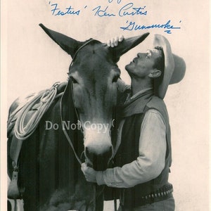 May include: A black and white photo of a man in a cowboy hat and vest, petting a donkey. The man is smiling and the donkey is looking at him. The photo is signed "Festus' Ken Curtis 'Gunsmoke' with a mess of good luck."