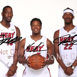 May include: Three basketball players wearing white jerseys with red accents and the word "HEAT" on the front. The players are standing in a row, with the player in the middle holding a basketball. The players' names and numbers are visible on their jerseys: "D. Wade 3", "K. Lowry 7", and "J. Butler 22".
