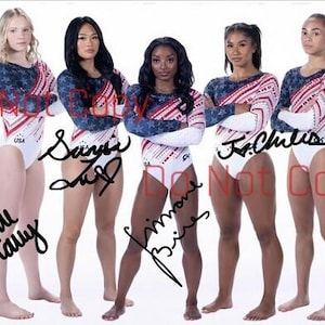 May include: Five female gymnasts wearing white and red leotards with stars and stripes. The leotards are long-sleeved and have a white base with red and blue stripes. The gymnasts are standing in a line, with their arms crossed in front of them. The gymnasts are all smiling and looking at the camera. The gymnasts are all wearing white socks and white shoes. The gymnasts are all standing on a white background. The gymnasts are all signed with their names.