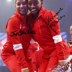 May include: Two women in red tracksuits with white accents, smiling at the camera. One tracksuit has "USA" printed on it. The image includes signatures. One is holding a bouquet of flowers.