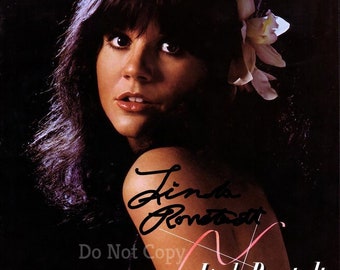 May include: A black and white photo of Linda Ronstadt, a singer, with long dark hair and a white flower in her hair. The photo is signed "Linda Ronstadt" in black ink.