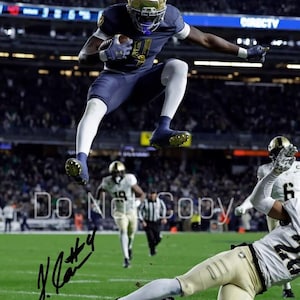 May include: A football player in a navy blue and gold uniform leaps in the air, holding a football. The player wears a gold helmet and white leggings. Other players in white and gold uniforms are visible on the field.